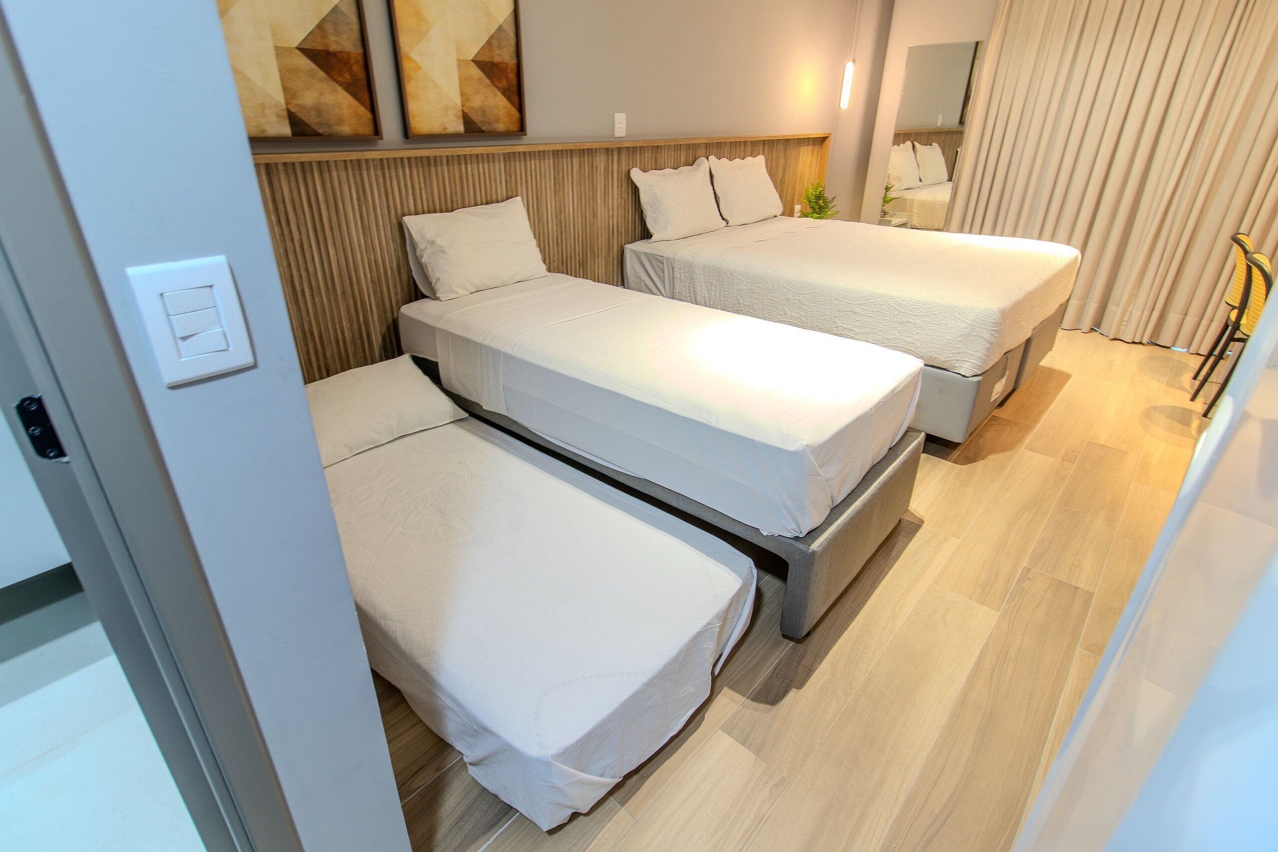 Une chambre d’hôtel avec trois lits, une tête de lit en bois, un miroir et une fenêtre avec des rideaux.