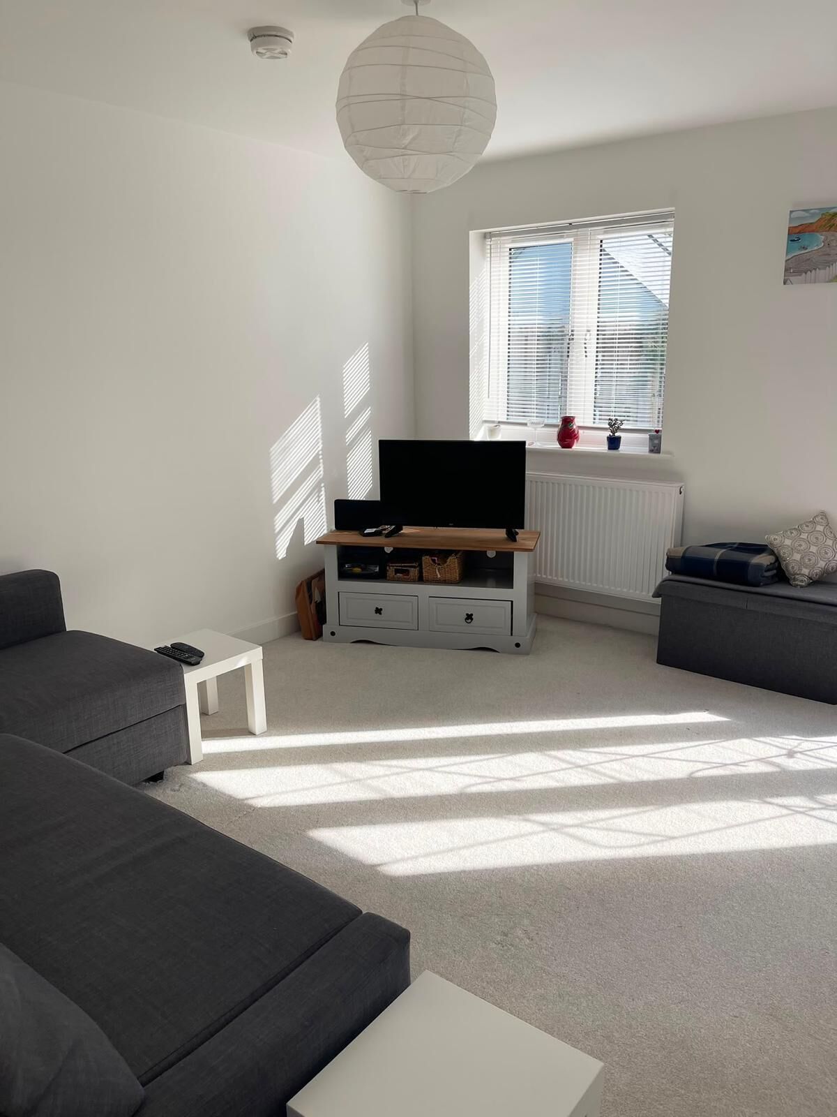A modern living room with a grey sofa, a white coffee table, a TV on a wooden stand, and a window with blinds.
