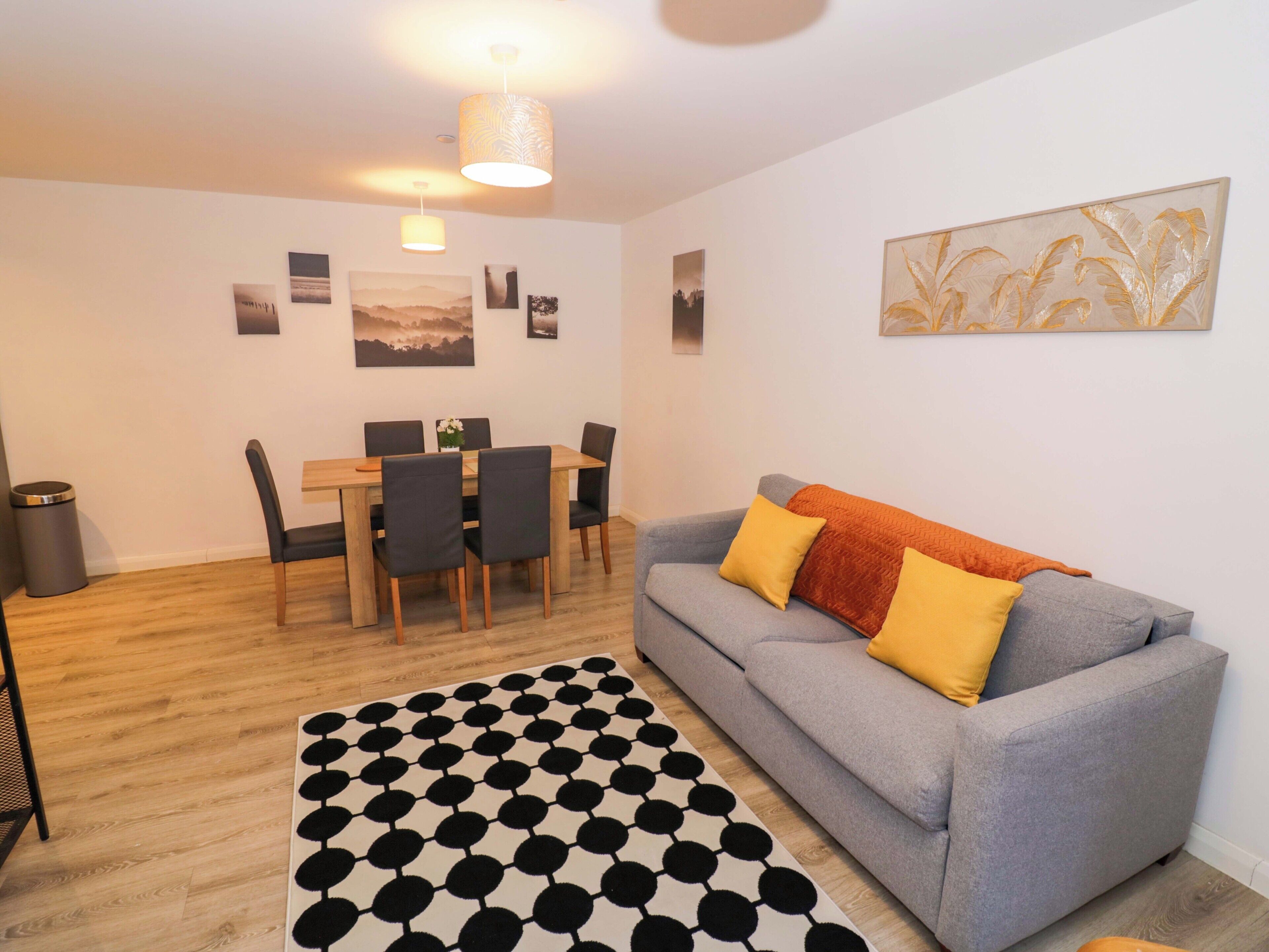 A living room with a grey sofa, a black and white patterned rug, a dining table with chairs, and wall art.