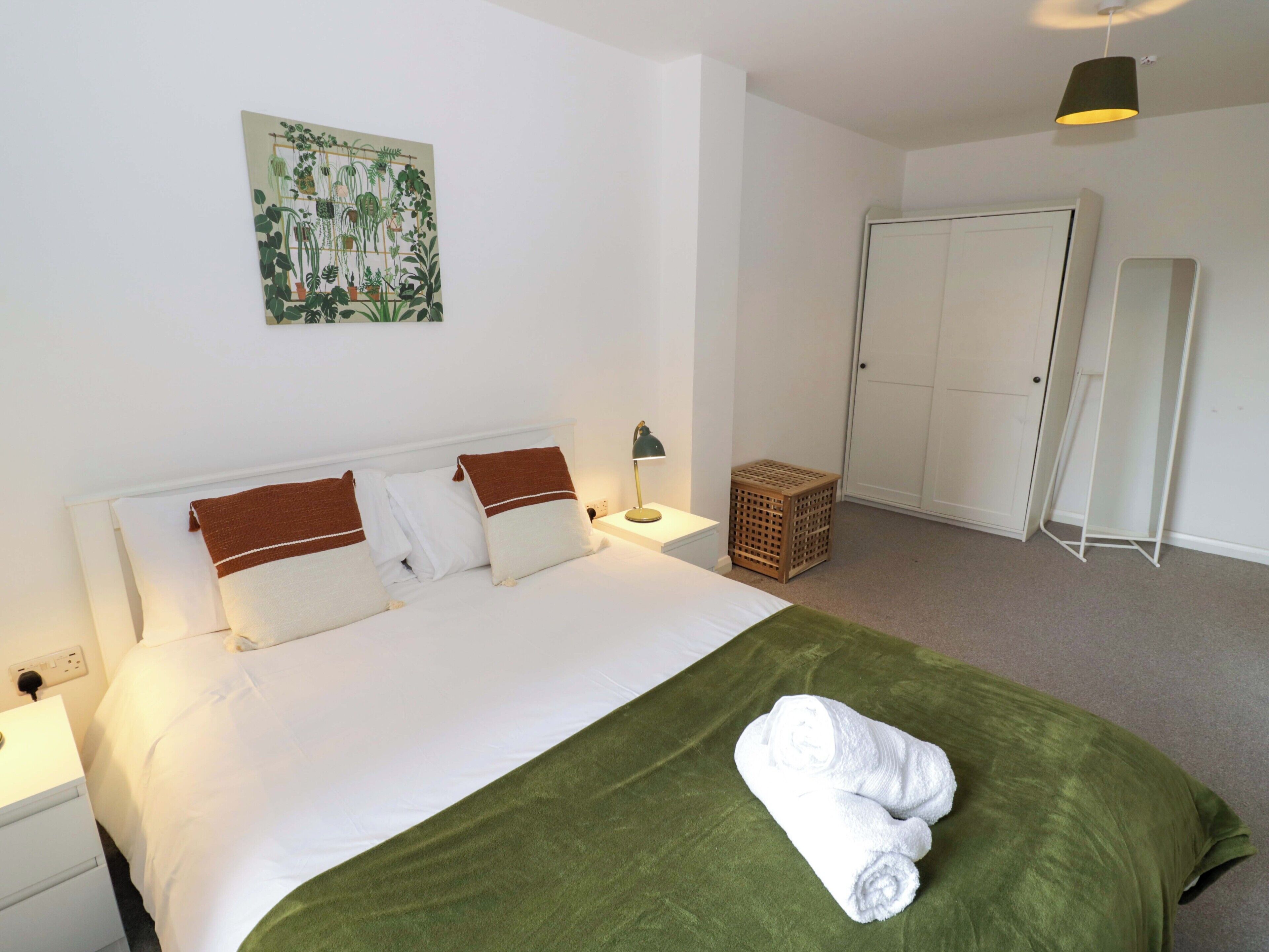 Een netjes opgemaakt bed met kussens, een groene deken en opgerolde handdoeken. Er staat een bedlampje, een klein houten mandje en een aan de muur bevestigde plant.