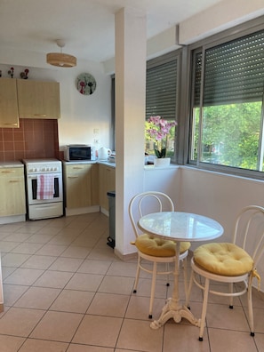 Une cuisine équipée d’une cuisinière, d’un four à micro-ondes et d’une fenêtre avec des stores. On y trouve une table avec deux chaises et un pot de fleurs posé sur le rebord de la fenêtre.