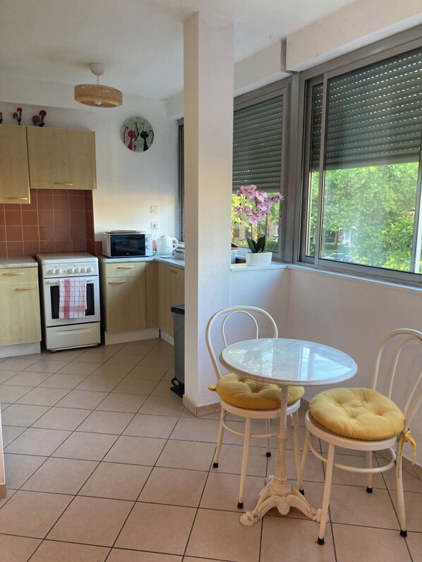 A kitchen with a stove, microwave, and a window with blinds. There is a table with two chairs and a potted plant on the windowsill.
