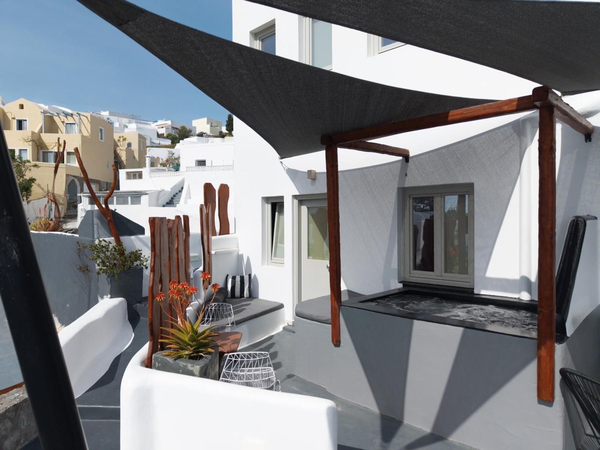 A rooftop terrace with a wooden pergola, white walls, and a view of neighboring buildings.