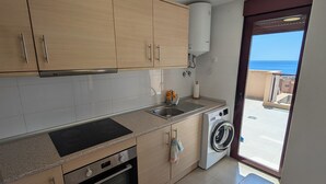 Cozinha com armários de madeira, forno embutido, máquina de lavar e vista para o mar através de uma porta.