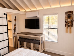 A bedroom with a bed, a desk with a chair, a TV, a window with blinds, and a wall-mounted artwork. - The Cabins at Indian Hills (Salome)