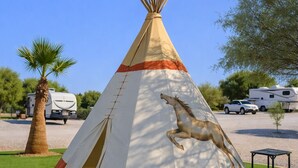 A teepee with a horse design, surrounded by grass and palm trees, with RVs in the background.