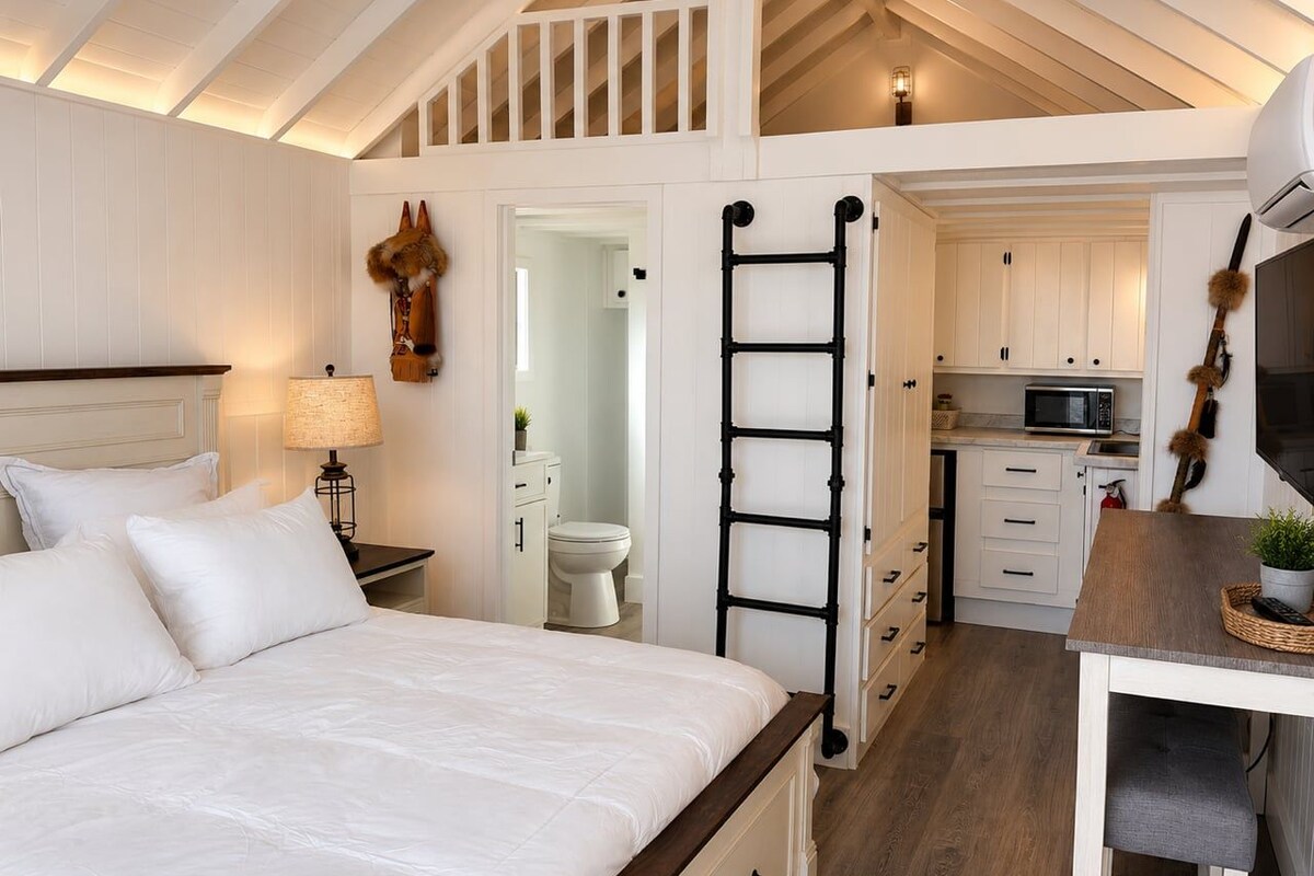 A bedroom with a bed, bedside table, lamp, and a view into the kitchen.
