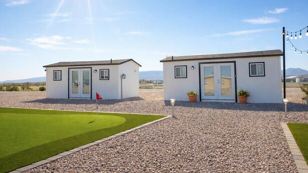 Two small, white cottages with doors and windows, surrounded by gravel and grass, with a clear sky and distant mountains.