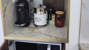 A kitchen with a microwave, coffee maker, kettle, and a shelf with glasses and a decorative plate.