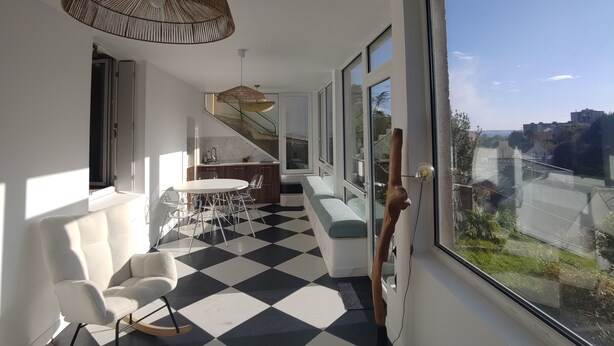 A modern interior with a checkered floor, a dining area, a staircase, and a large window offering a city view.
