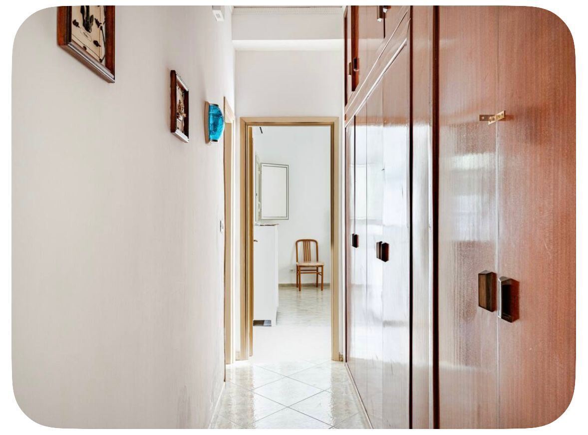 Un corridoio con una porta in legno, una sedia e un muro bianco decorato con quadri.