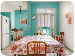 A dining area with a table covered in a floral tablecloth, wooden chairs, a white refrigerator, a clock on the wall, and a window with curtains. - SeaHouse Palermo| Authentic relaxation near Mondello and historic center (Palermo)