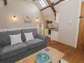 A cozy room with a grey sofa, a wooden table with a blue glass bowl, a kitchen in the background, and a sloped ceiling with exposed beams. - Beudy Bach (Llanbedr)