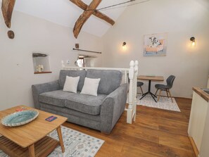 A cozy room with a grey sofa, a wooden table, a small desk, a chair, and a bed with white headboard. - Beudy Bach (Llanbedr)