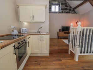 A compact kitchen with a stove, oven, and white cabinets. A dining area with a TV and wooden furniture is visible. - Beudy Bach (Llanbedr)