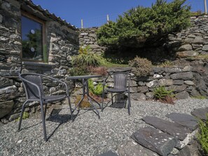 A stone building with a window, two wicker chairs, a small table, and a garden with shrubs and a tree. - Beudy Bach (Llanbedr)