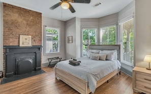 A bedroom with a fireplace, a bed with striped bedding, a ceiling fan, and large windows with blinds. - Charming 2-bedroom, 2 bath w/ Porches • Walk to Forsyth Park (Savannah)