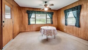 Une pièce aux murs en bois, avec un ventilateur au plafond, une table ronde recouverte d’une nappe à motifs floraux et deux fenêtres ornées de rideaux à motifs.