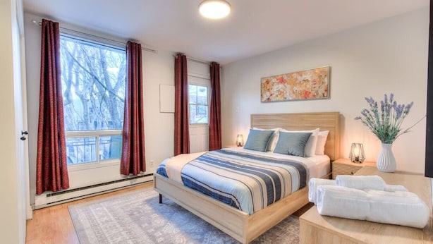 A bedroom with a bed, bedside tables, a nightstand, a window with curtains, a painting on the wall, and a vase with flowers. - Stylish Plateau 1BR near Parc La Fontaine (Montreal)