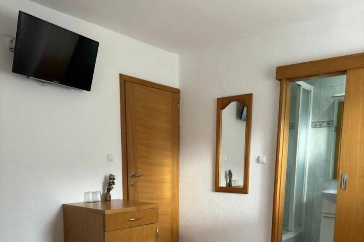 A hotel room with a bed, a wooden wardrobe, a television, a mirror, and a bathroom door.