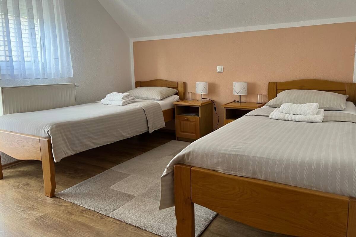 A hotel room with two single beds, a wooden headboard, bedside tables with lamps, a window with blinds, and a carpeted floor.