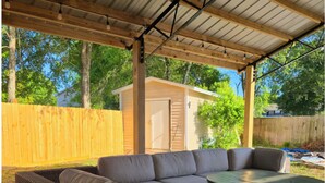 Un patio exterior con un sofá de mimbre, una mesa redonda y una pérgola adornada con luces colgantes.
