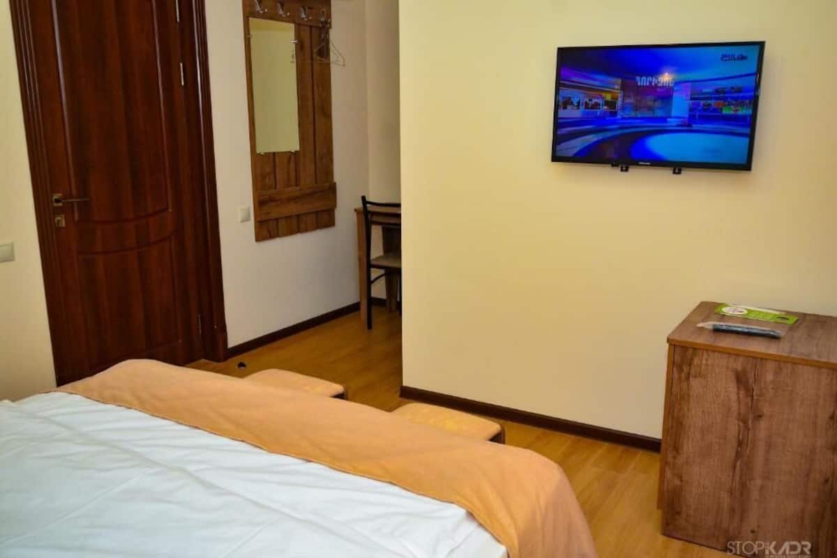A hotel room with a bed, a wooden wardrobe, a television mounted on the wall, and a small wooden cabinet.