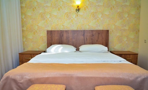 Une chambre d’hôtel avec une tête de lit en bois, deux tables de chevet, un lit avec du linge de lit blanc et un papier peint à motifs floraux.