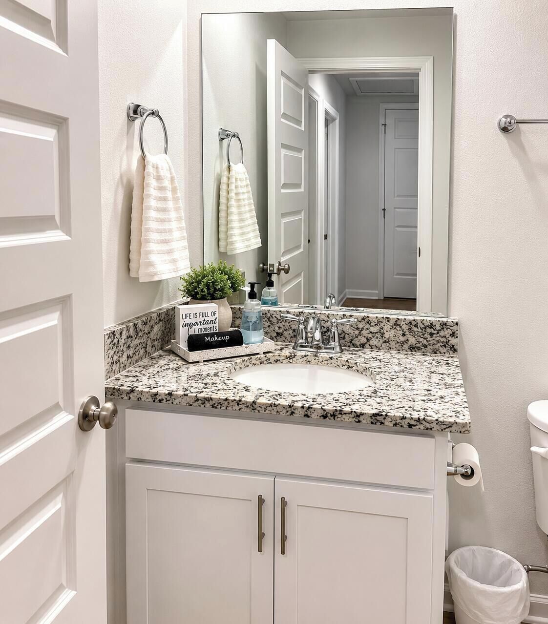 A bathroom with a granite countertop, white cabinets, a mirror, a towel rack with towels, a small plant, and a makeup station.