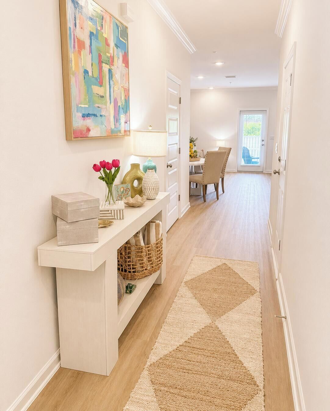 A hallway with a white console table, a colorful abstract painting, a door leading to a dining area, and a patterned rug.