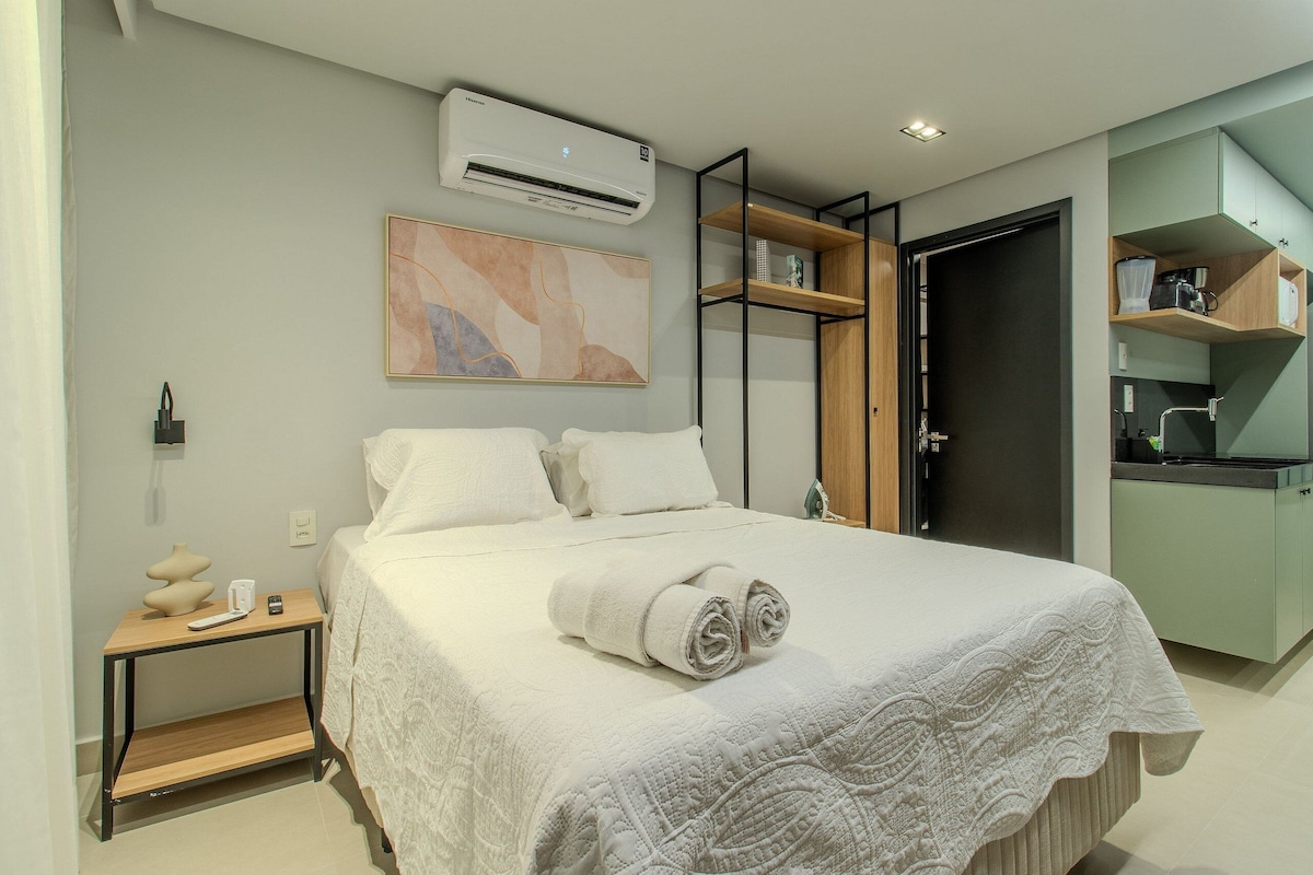 A modern bedroom with a bed, bedside table, air conditioning unit, and a kitchenette area.