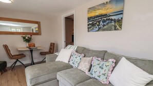 A living room with a grey sectional sofa, a small dining table with two chairs, a mirror, and a painting of a coastal scene. - Little Holm Garth (Whitby)