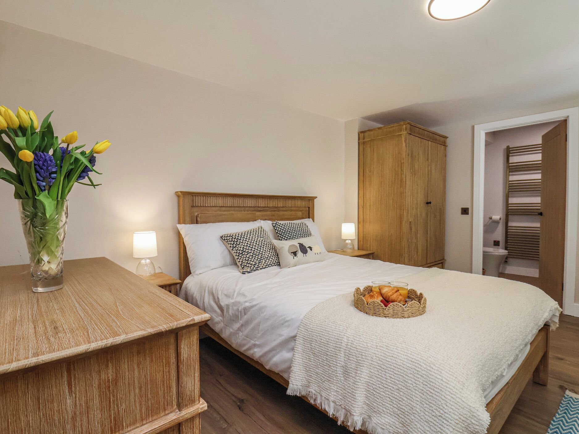 A bedroom with a wooden headboard, a bedside table with a vase of flowers, a bed with white bedding, a basket of bread, and a wardrobe.