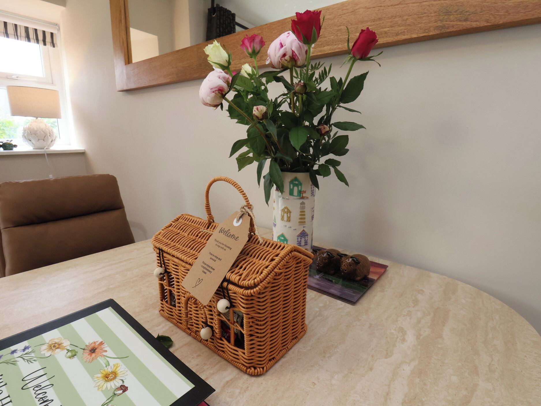 Ein Korbkörbchen mit einem Anhänger, eine Vase mit Rosen und ein gerahmtes Willkommensschild auf einer Tischplatte.