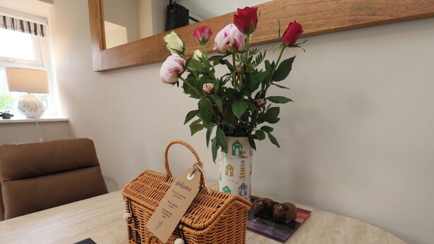 A wicker basket with a tag, a vase with roses, and a framed welcome sign on a table. - Little Holm Garth (Whitby)