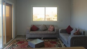 A living room with a grey sofa set, a patterned rug, a ceiling light fixture, and a large window with a view of buildings.