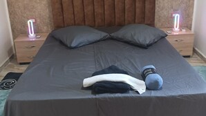 A neatly made bed with a grey bedspread, two pillows, and a few folded clothes on the bed. There are two bedside tables with illuminated signs, a brown upholstered headboard, and a textured wall behind the bed.