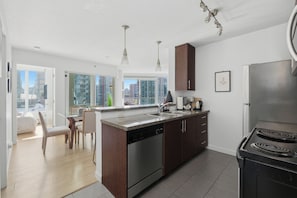 A modern kitchen with a dishwasher, stove, and dining area with chairs and a table. - Sky-High Luxury 2BR 2BA | Panoramic View + DT Core (Vancouver)