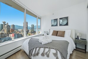 A hotel room with a large window, a bed, two wall-mounted artworks, and a nightstand with a lamp. - Sky-High Luxury 2BR 2BA | Panoramic View + DT Core (Vancouver)