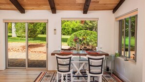 Un coin repas avec une table dressée pour deux personnes, deux chaises, un tapis et une vue sur un jardin, visible à travers de grandes fenêtres.