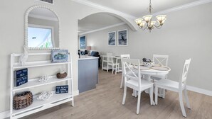 Ein Essbereich mit einem weißen Tisch, Stühlen und einer Deckenleuchte. Es gibt eine Küchenzeile mit einem Spülbecken und einem Fenster.