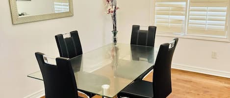 A dining area with a glass table, black chairs, a vase with flowers, a mirror, and a window with blinds.
