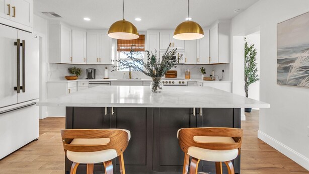 A modern kitchen with white cabinets, a large island, two bar stools, and pendant lighting. - Palms of the Isles (St. Petersburg)