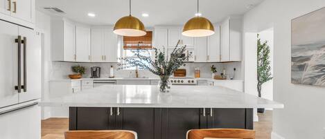 Una cocina moderna con armarios blancos, una isla grande, dos taburetes de bar e iluminación colgante.