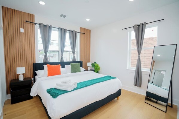 A modern hotel room with a large bed, a nightstand with a lamp, a mirror, and a view of the outside. - Modern Home Near MetLife | 2BR | Free Parking (Jersey City)