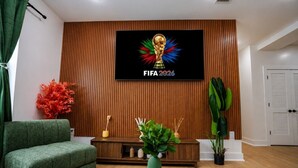 Moderni olohuone, jonka seinällä on televisio, jossa näkyy peli «FIFA 2026». Huoneessa on vihreä sohva, puinen konsolipöytä koriste-esineineen ja suuri huonekasvi.