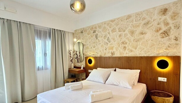 A hotel room with a large bed, a stone wall, a round mirror, and a wooden nightstand. - Kapetánissa Concept Studios (Stalida)