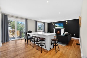 A modern living room with a central island, bar stools, a fireplace, and a view of the outdoors.