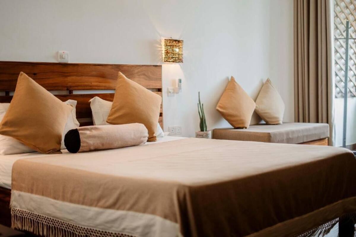 A bedroom with a wooden headboard, two beds with beige bedding, a wall-mounted light fixture, and a potted plant.