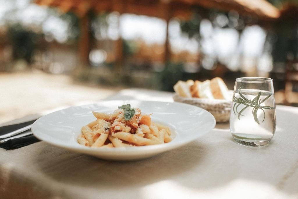 A plate of pasta with a glass of water and a basket of bread on a table.